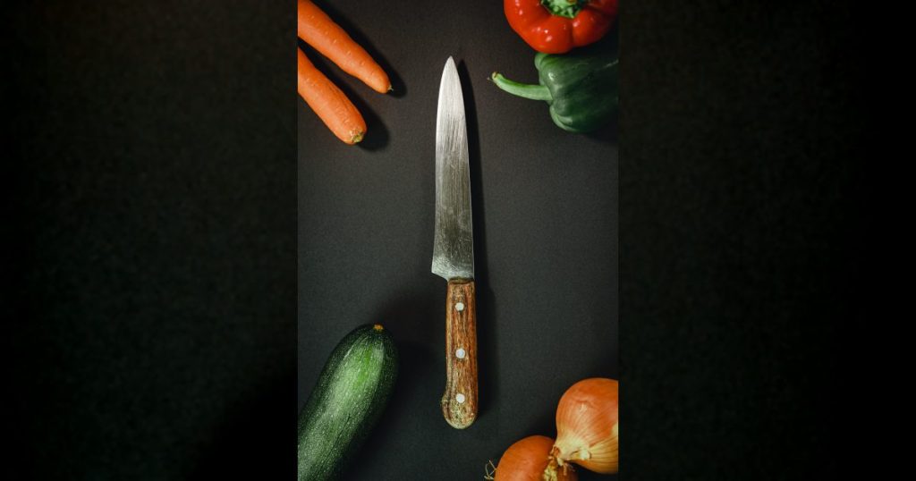 Chef knife surrounded by fresh vegetables on a dark work surface